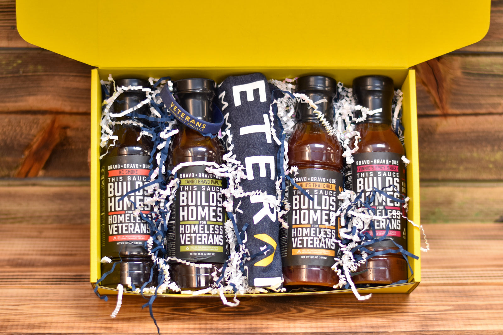 A gift box open to reveal multiple bottles of barbecue sauce and a t-shirt with 'Veterans Community Project' branding, all arranged on a wooden surface.