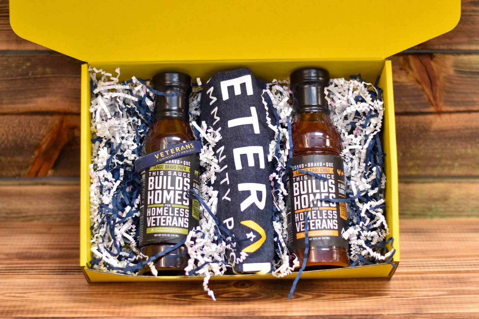 A gift box open to reveal two bottles of barbecue sauce, a bracelet, and a classic T-shirt inside, all arranged on a bed of decorative shaving. The box and its contents have branding elements suggesting a collaboration with Veterans Community Project.