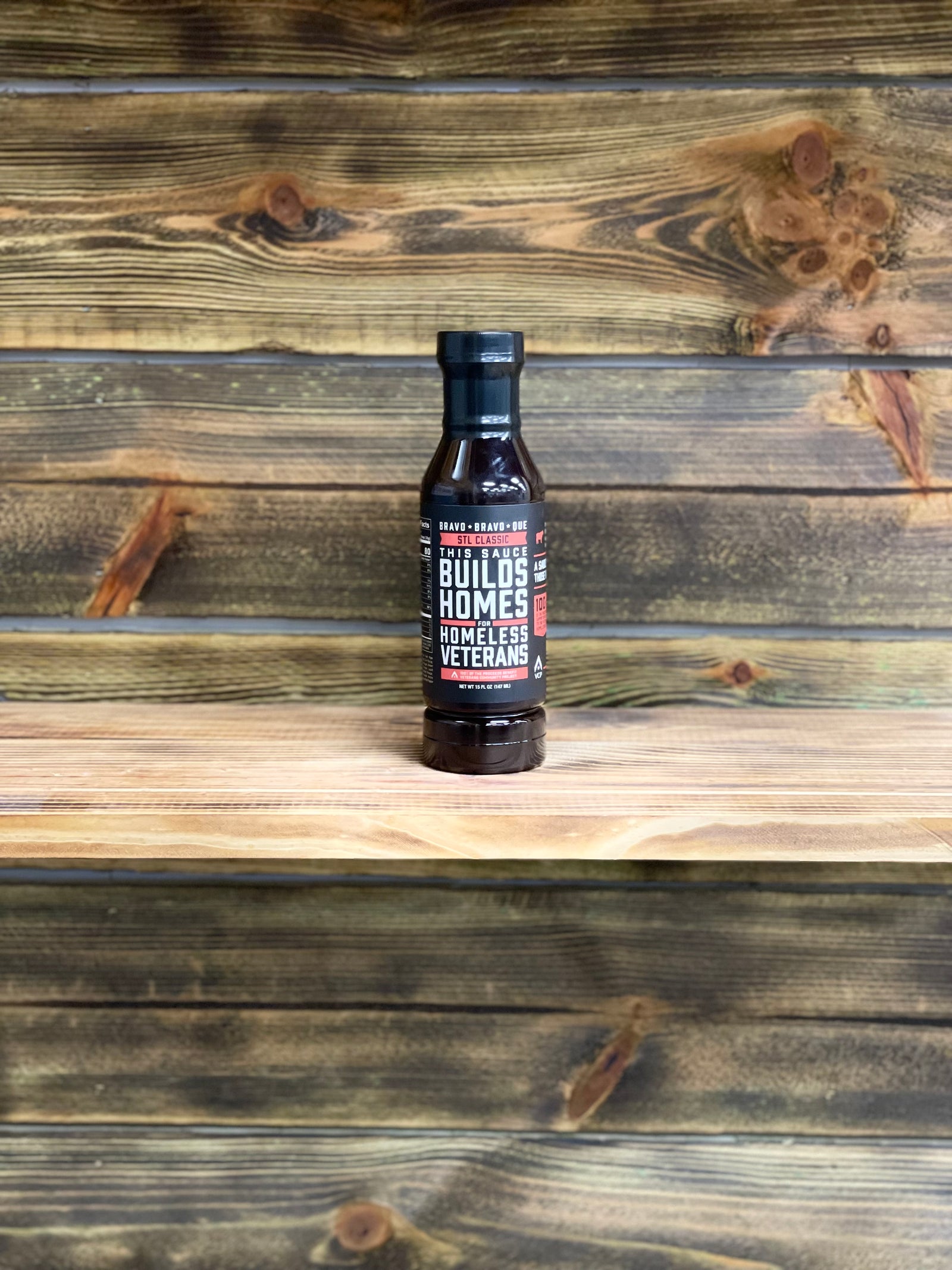 A bottle of 'Bravo Bravo Que's' STL Classic barbeque sauce placed on a wooden surface with a rustic background.
